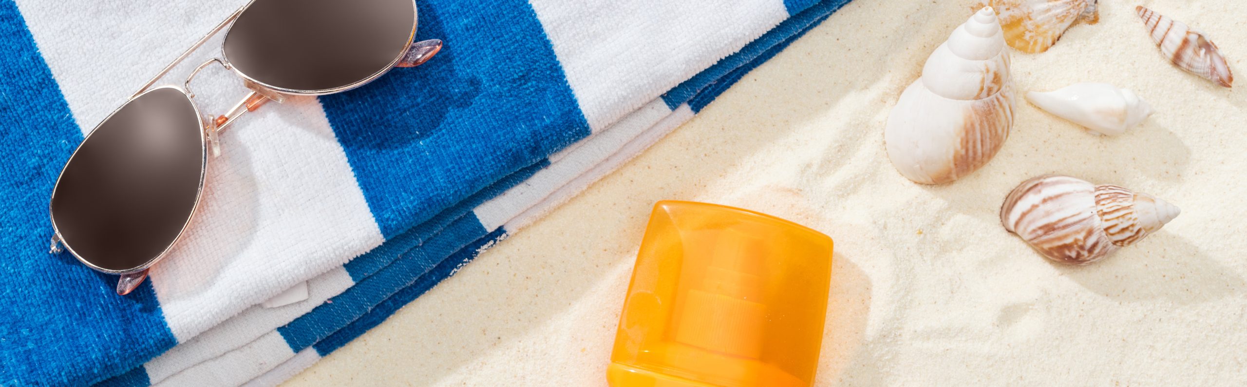 orange bottle of sunscreen on sand with seashells, striped towel and sunglasses, panoramic shot